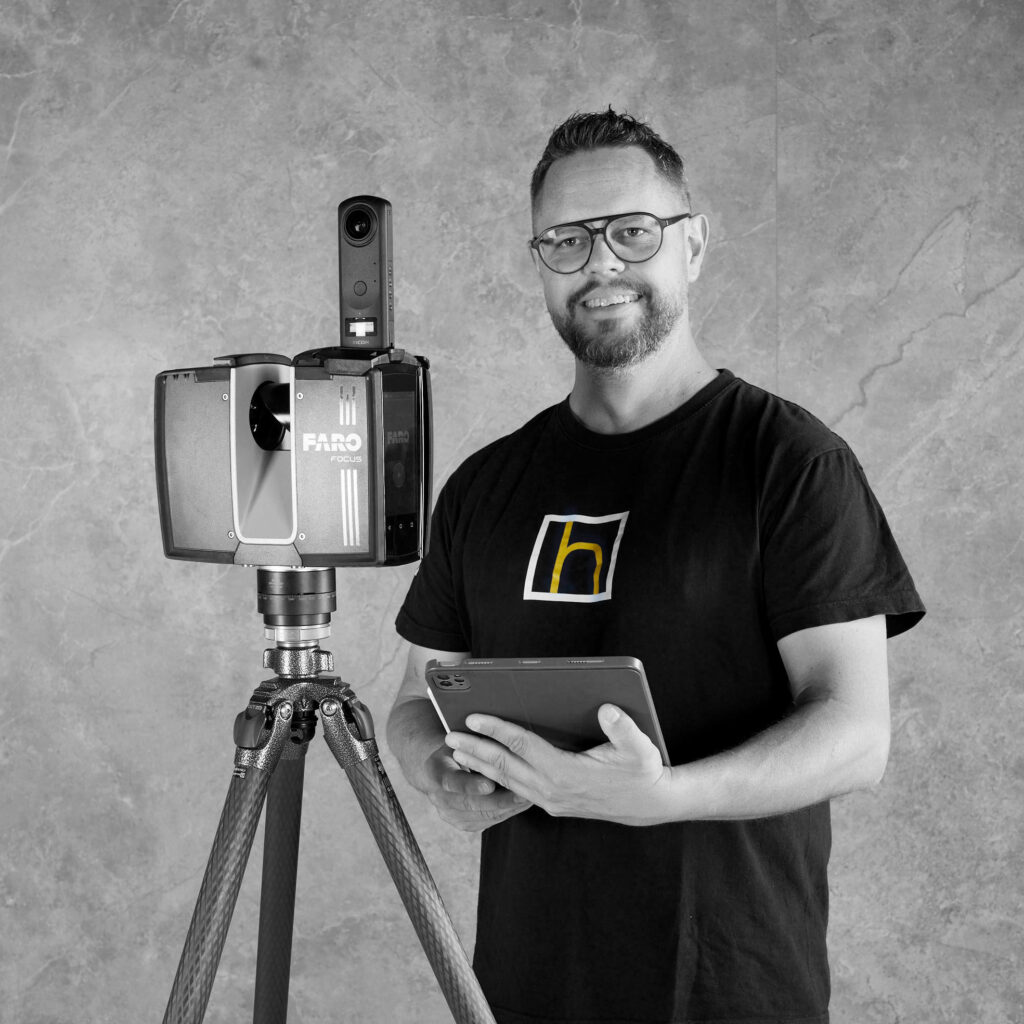 Ein lächelnder Mann mit Bart hält ein Tablet neben einer Kamera auf einem Stativ.